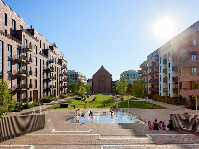 Weitwinkelaufnahme eines zentralen Quartiersplatzes mit Wasserspiel, flankiert von modernen Wohngebäuden und offener Grünachse bis zum historischen Bauwerk im Hintergrund.