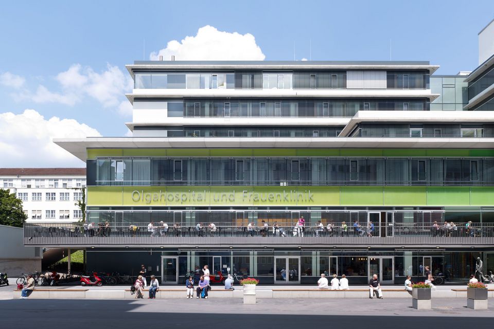 Außenansicht eines Klinikgebäudes mit grüner Fassadenverkleidung und breiter Terrasse, klare horizontale Gliederung