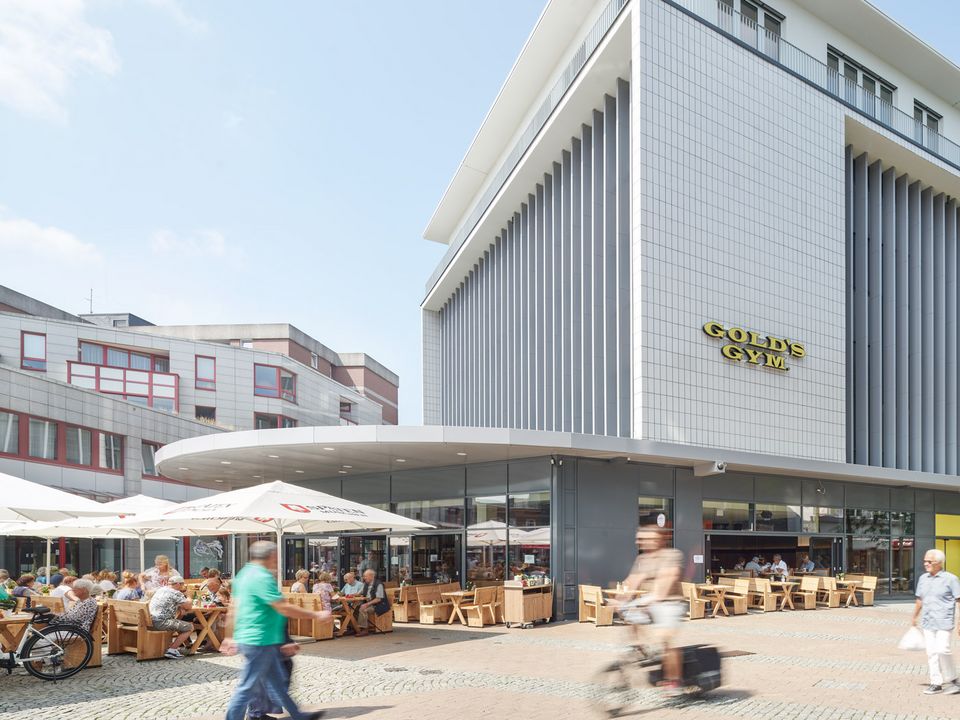 Außenansicht eines Gebäudes mit vertikalen Fassadenelementen und Glasfront, davor Außengastronomie mit Sonnenschirmen und Sitzgruppen