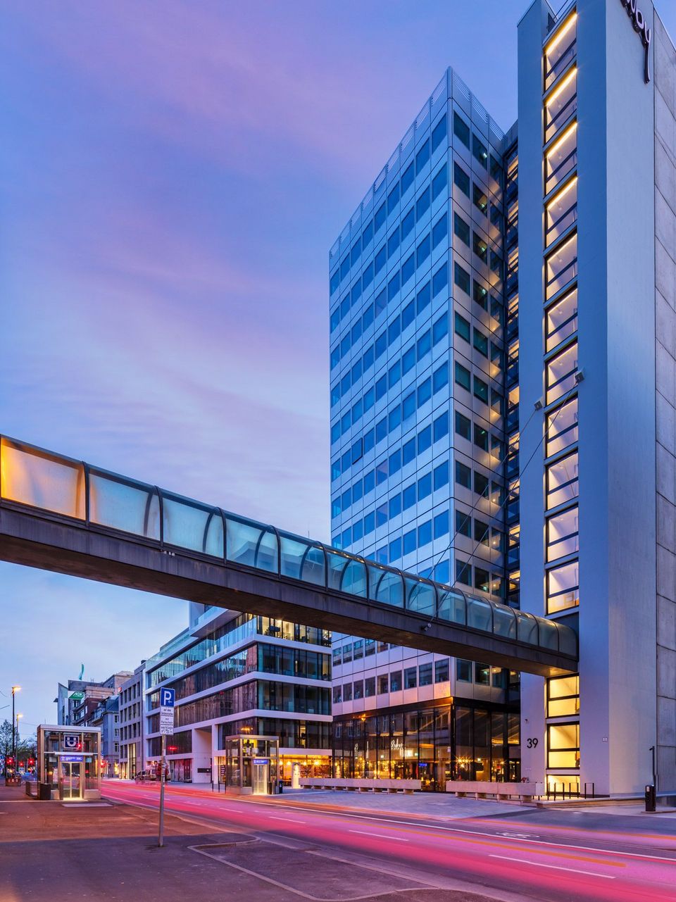 Straßenansicht eines modernen Hochhauses mit Glas- und Metallfassade, Fußgängerbrücke verbindet Gebäude, abendliche Beleuchtung