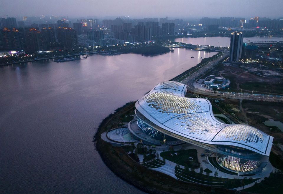 Luftaufnahme eines skulpturalen Gebäudekomplexes am Wasser mit weitläufiger Dachlandschaft, geschwungenen Außenräumen und angrenzender Uferpromenade.