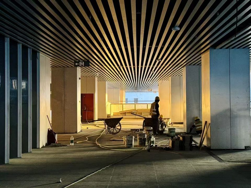 Innenaufnahme einer großzügigen Lobby im Ausbauzustand mit linearem Deckenraster, sichtbaren Installationen und arbeitenden Baukräften im Vordergrund.