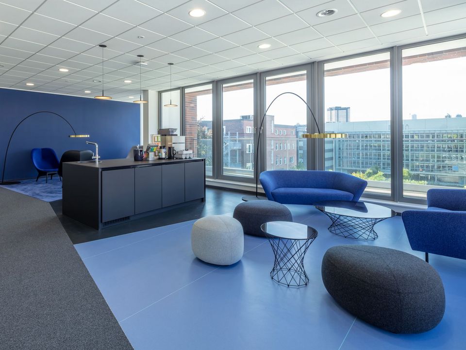 Innenaufnahme einer offenen Loungezone mit Teeküche, Sitzlandschaften und raumhoher Verglasung mit Blick in den Campus.