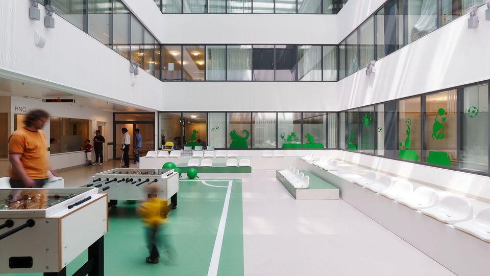 Innenhof einer Klinik mit Spielfläche in Grün und Weiß, Tischkicker und Sitzbänken, umlaufende Glasfronten und Oberlicht