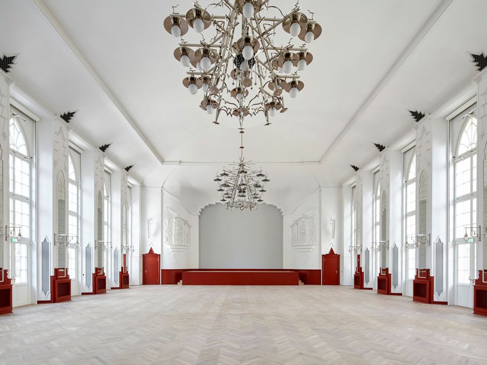 Innenaufnahme eines großen Veranstaltungssaals mit hohen Fenstern, dekorativen Wandreliefs und mehrarmigen Kronleuchtern, die den repräsentativen Charakter des Raumes hervorheben.