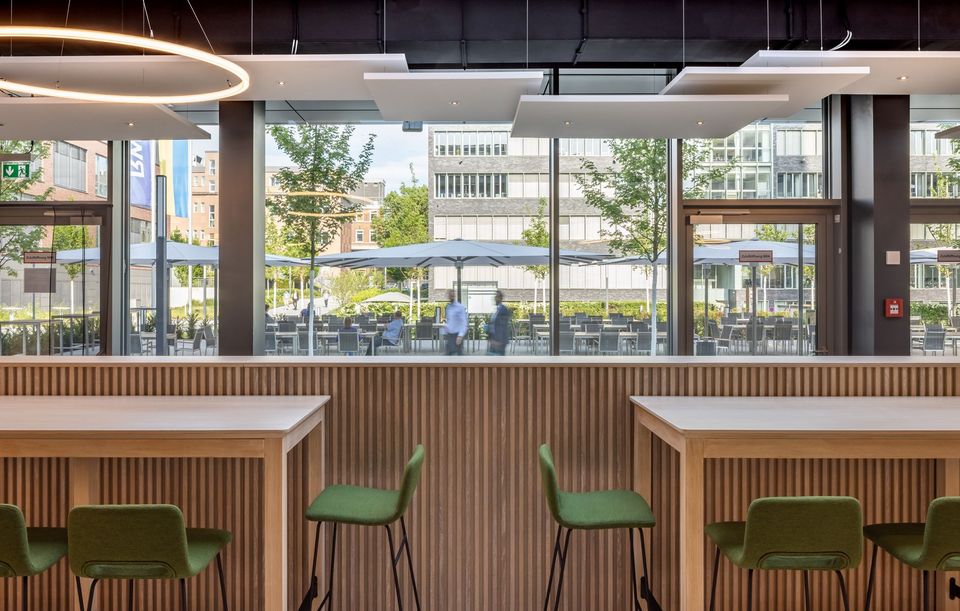 Innenaufnahme einer Café- und Aufenthaltszone mit Hochtischen, Akustikpaneelen und großformatiger Fensterfront mit Blick in den Außenbereich.