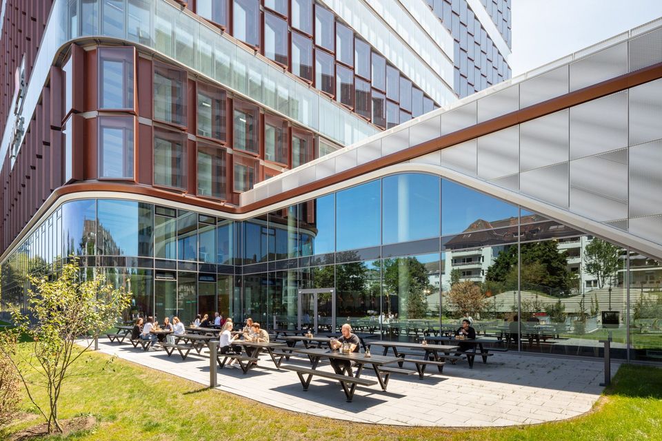 Außenbereich mit Sitzbänken vor verglaster Fassade eines Bürogebäudes, markante Architektur mit roten Fassadenelementen