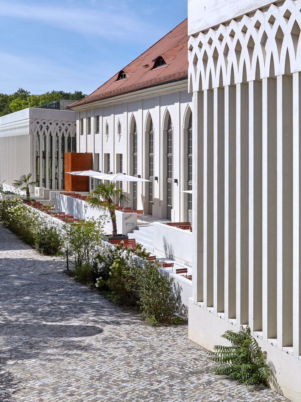Außenansicht eines langgestreckten Gebäudekörpers mit vertikaler Pfeilerfassade, Bogenfenstern und vorgelagertem Gartenweg, der die Verbindung von Architektur und Landschaft zeigt.