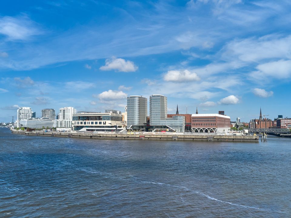 Außenaufnahme einer städtischen Platzsituation mit gemischt genutzten Gebäuden, markanten Arkaden und einem modernen Hochhausensemble entlang der Uferpromenade