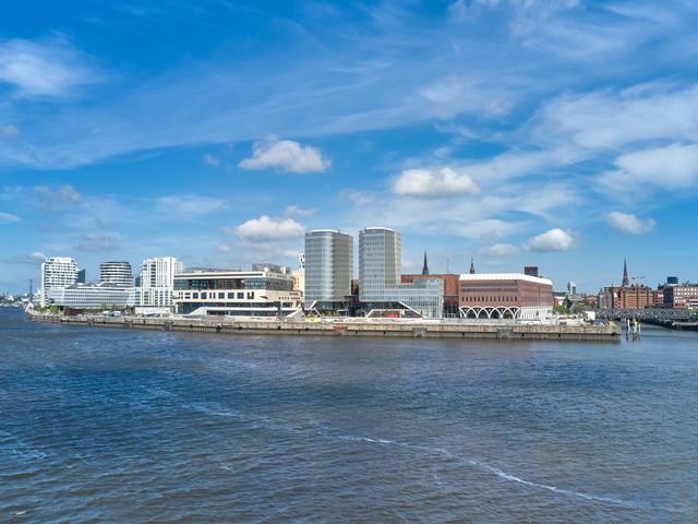 Außenaufnahme einer städtischen Platzsituation mit gemischt genutzten Gebäuden, markanten Arkaden und einem modernen Hochhausensemble entlang der Uferpromenade
