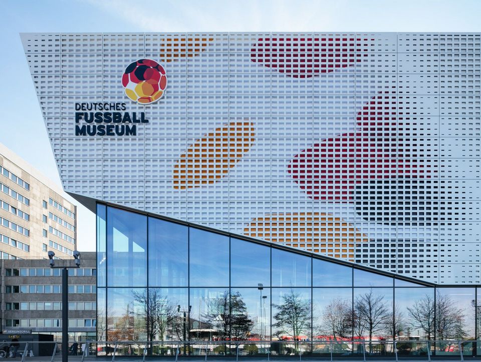 Außenaufnahme der Museumsfassade mit großformatigem Logo, farbigen Fassadenfeldern und transparentem Eingangsbereich, die die ikonografische Architektur des Deutschen Fußballmuseums zeigt.