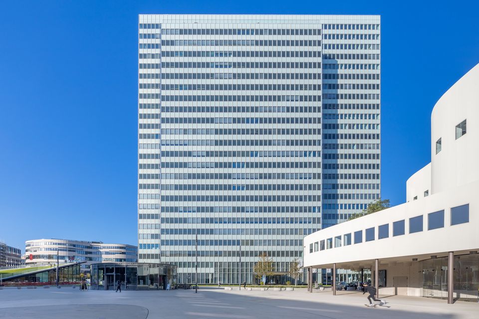 Modernes Hochhaus mit klarer Rasterfassade aus Glas und Aluminium, umgeben von offenen Platzflächen und angrenzenden Gebäuden