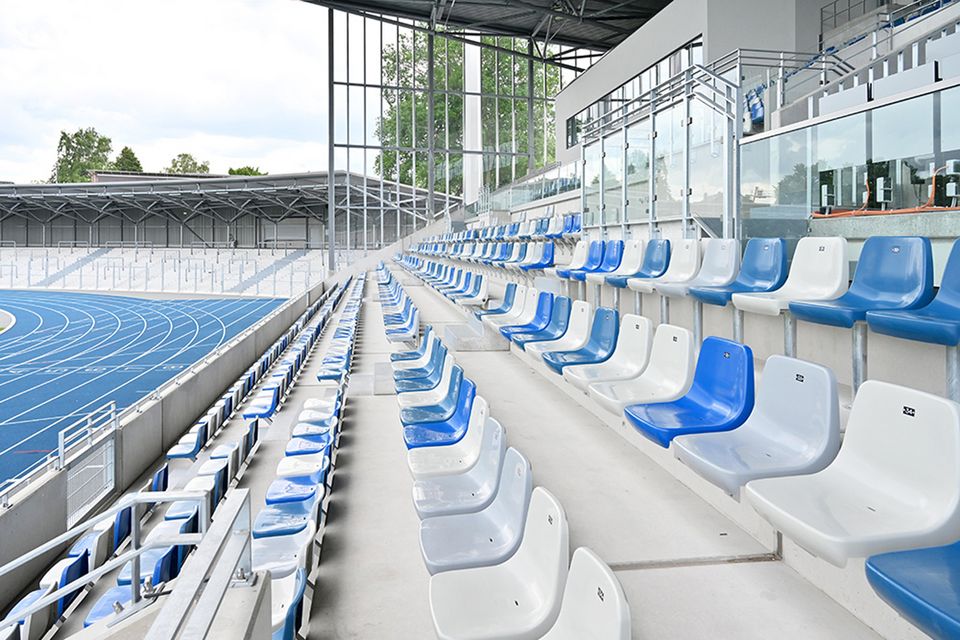 Innenaufnahme eines Tribünenbereichs mit farblich abgesetzten Sitzschalen, verglaster Stirnseite und Blick auf die Laufbahn.