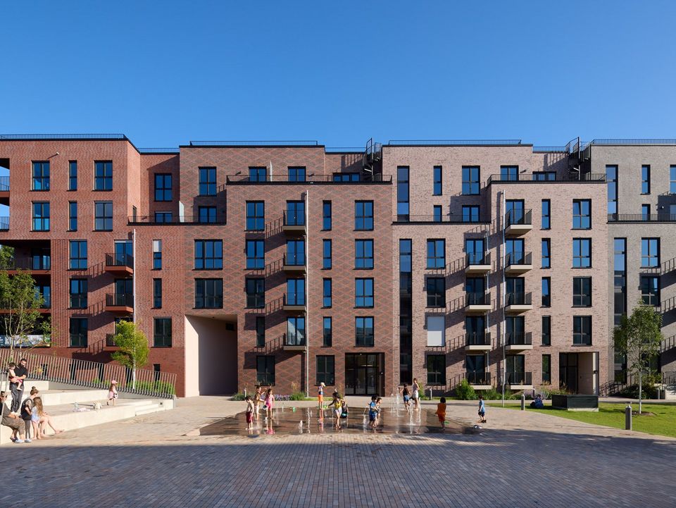 Frontalaufnahme eines Wohngebäudes mit rhythmisierter Klinkerfassade, Balkonzonen und belebtem Quartiersplatz mit Wasserspiel.