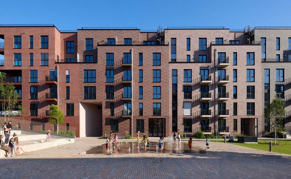 Frontalaufnahme eines Wohngebäudes mit rhythmisierter Klinkerfassade, Balkonzonen und belebtem Quartiersplatz mit Wasserspiel.