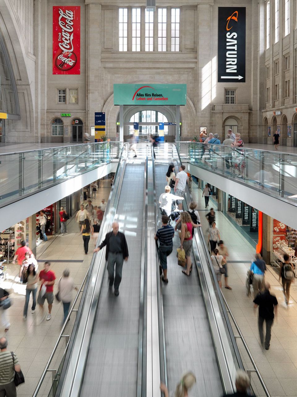 Frontalansicht einer breiten Rolltreppenanlage im Hauptbahnhof mit oberer Eingangshalle, Retail‑Geschäften auf mehreren Ebenen und architektonisch klar gegliederten Wand- und Fensteröffnungen.