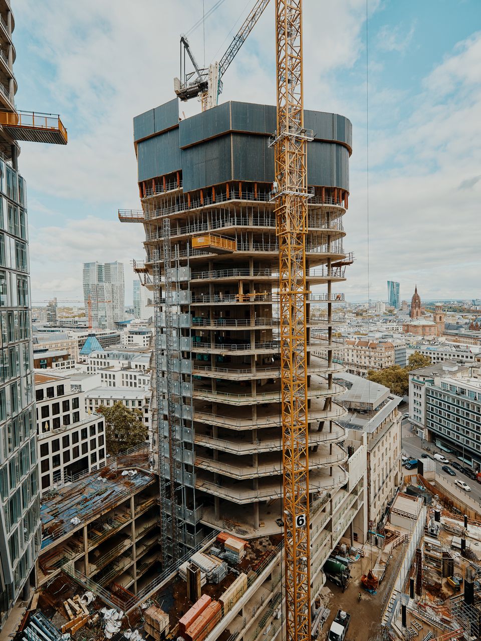 Weitwinkelaufnahme eines runden Hochhausrohbaus im oberen Ausbau mit umlaufenden Gerüsten, Turmdrehkran und städtischem Panorama im Hintergrund.