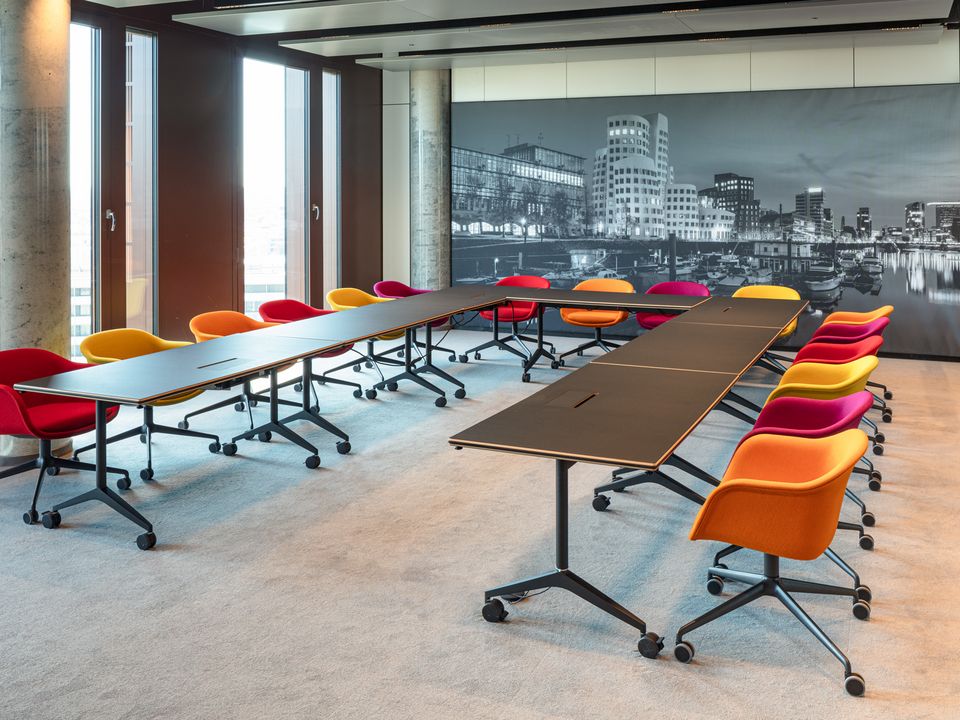 Innenaufnahme eines flexibel möblierbaren Konferenzraums mit U‑förmig angeordneten Tischen, farblich akzentuierten Drehstühlen und wandfüllender Stadtpanoramografie.
