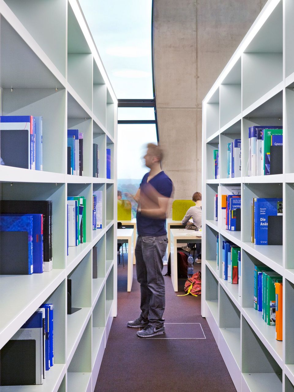 Bibliotheksgang mit hohen weißen Regalen voller Fachbücher, dahinter Arbeitsplätze vor großem Fenster