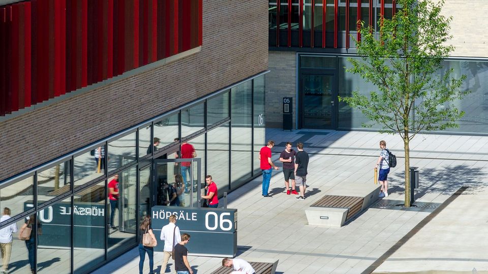 Außenansicht eines Hochschulgebäudes mit Eingangsbereich und Vorplatz. Studierende halten sich im öffentlichen Raum auf.