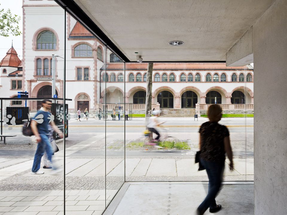 Durchblick von modernem Glasfoyer auf historisches Gebäude mit Arkaden und roten Ziegeldetails, Kontrast von Parkhaus und Altbau