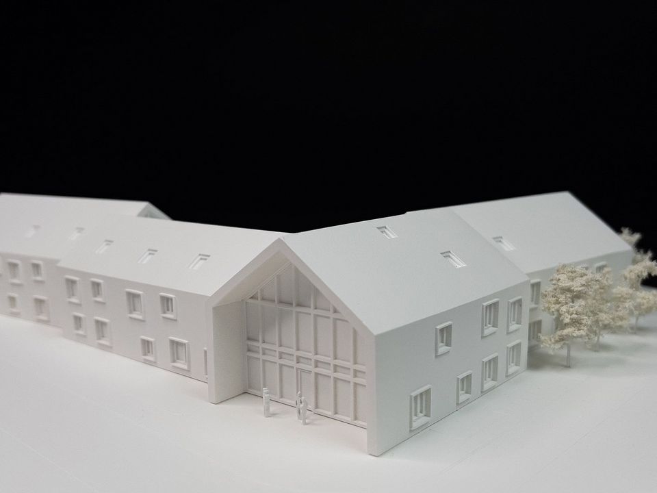 Modellaufnahme eines Gebäudes mit giebelständigem Haupteingang, klarer Fassadenstruktur und angedeuteten Außenanlagen in weißer Darstellung.