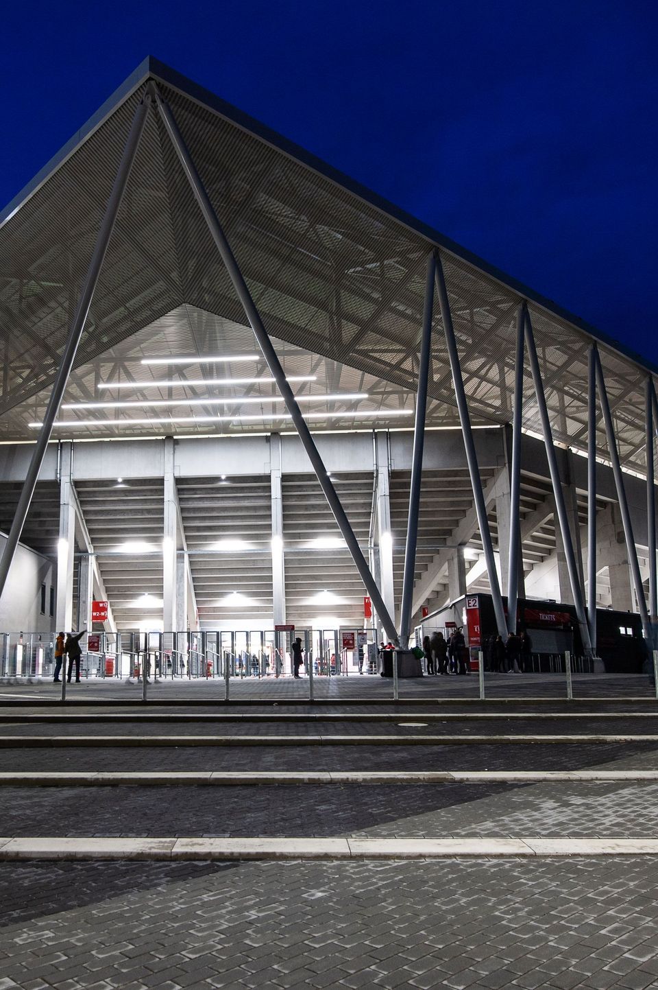 Abendansicht des Stadioneingangs mit beleuchteter Dachkonstruktion und schrägen Stahlstützen, moderne Architektur mit klarer Linienführung