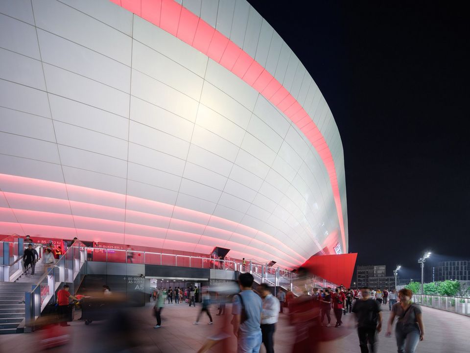 Außenaufnahme eines Fußballstadions bei Nacht mit leuchtender Stadionhülle, umlaufenden LED‑Bändern, breiten Zugängen und belebtem Vorplatz als Teil der Event‑Infrastruktur.