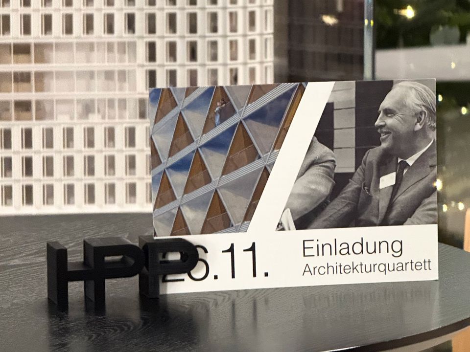 Detailaufnahme einer Einladungskarte zum Architekturquartett mit grafischen Fassadenelementen und HPP‑Schriftzug, platziert auf einer dunklen Tischoberfläche.