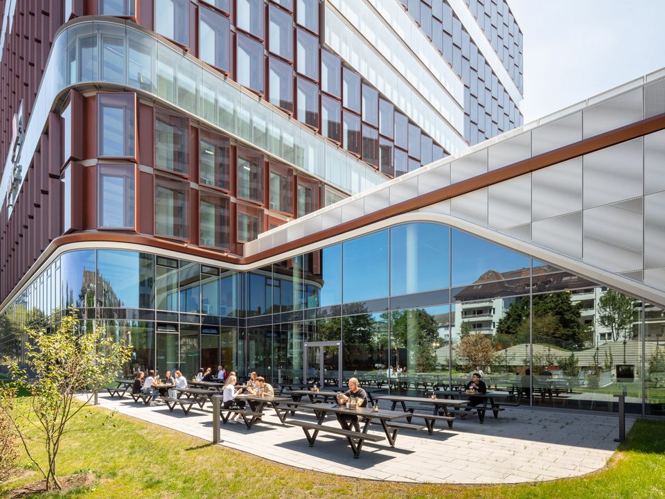 Außenbereich mit Sitzbänken vor verglaster Sockelfassade eines Bürogebäudes, markante Architektur mit roten Fassadenelementen