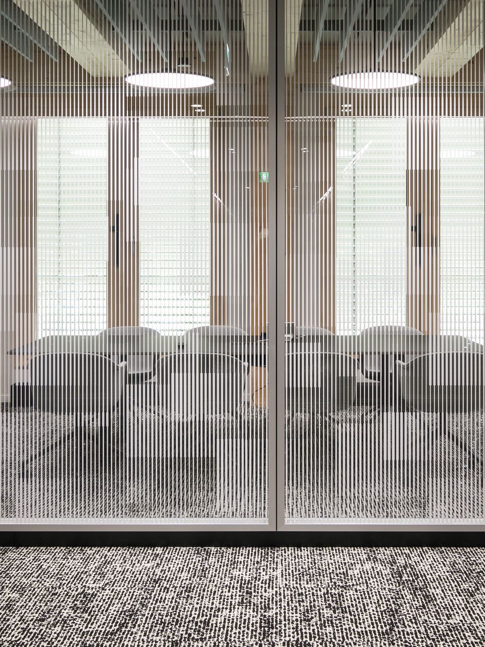 Besprechungsraum mit Glaswand und vertikalen Linien, dahinter moderne Sitzmöbel und strukturierter Teppichboden