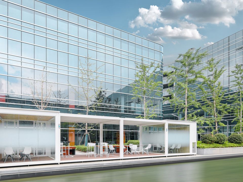 Außenaufnahme eines modernen Bürogebäudes mit verglaster Fassade und vorgelagerter Terrasse direkt am Wasserlauf.