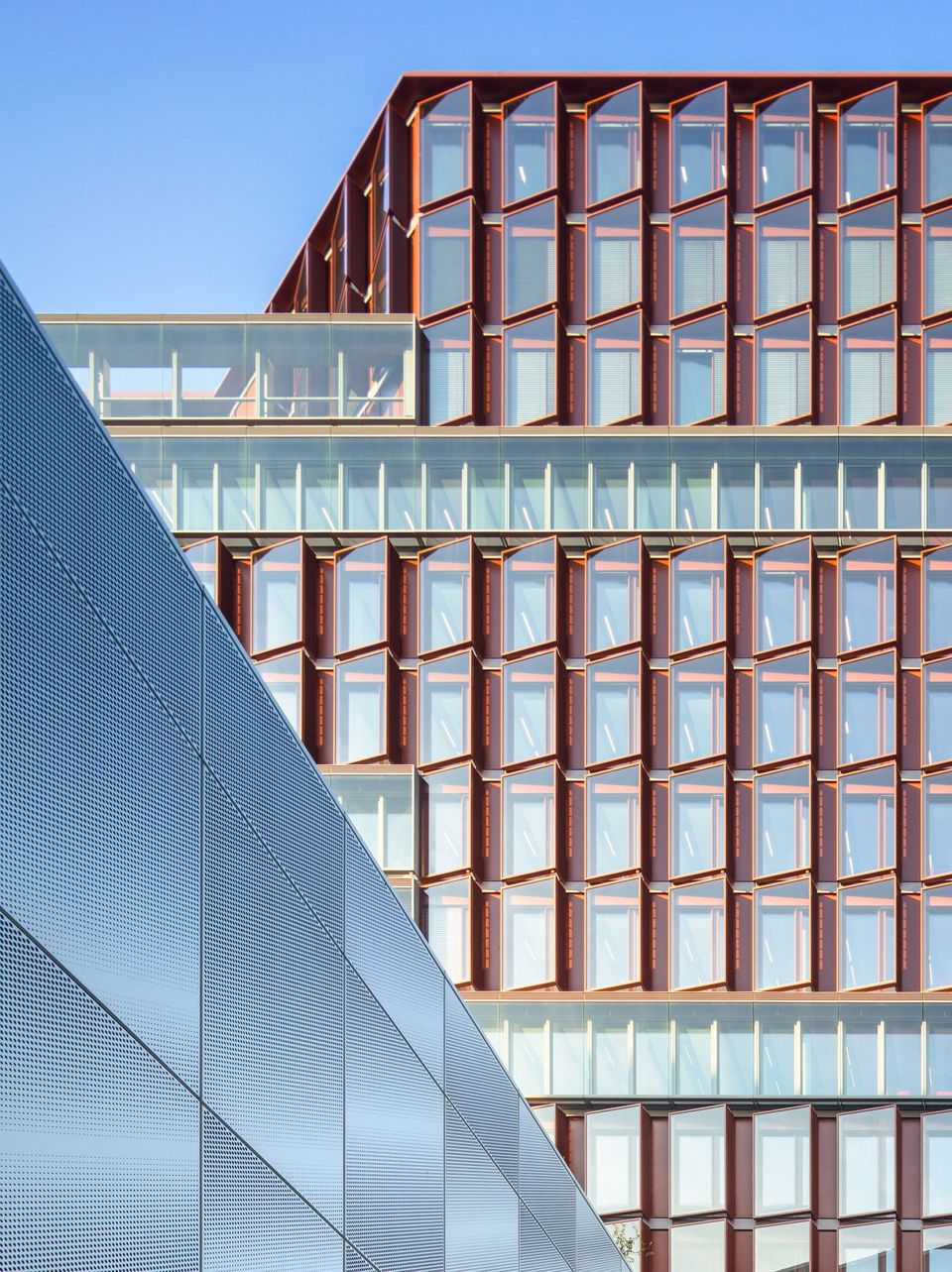 Detailansicht einer Glasfassade mit roten Fassadenprofilen und perforierter Metallverkleidung, klare Linien vor blauem Himmel