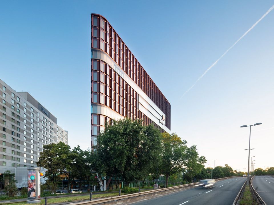 Straßenansicht eines markanten Bürogebäudes mit geschwungener Glasfassade und roten Fassadenelementen, flankiert von Bäumen und breiter Fahrbahn