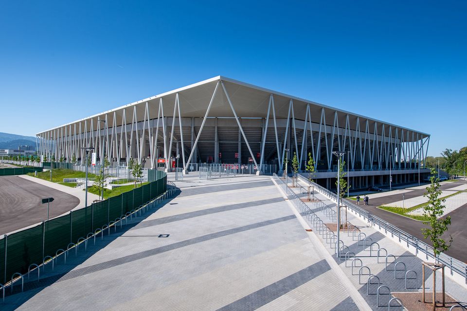 Außenaufnahme eines modernen Stadions mit markanter Stahlstützenstruktur, weit auskragendem Dach und offenem Vorplatzbereich im urbanen Umfeld.