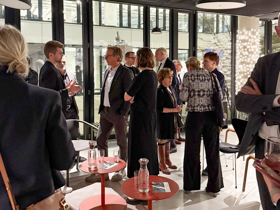 Übersichtsaufnahme eines Get‑togethers mit mehreren Personen in einem verglasten Innenraum mit Sitzgruppen, Stehtischen und abendlicher Atmosphäre.