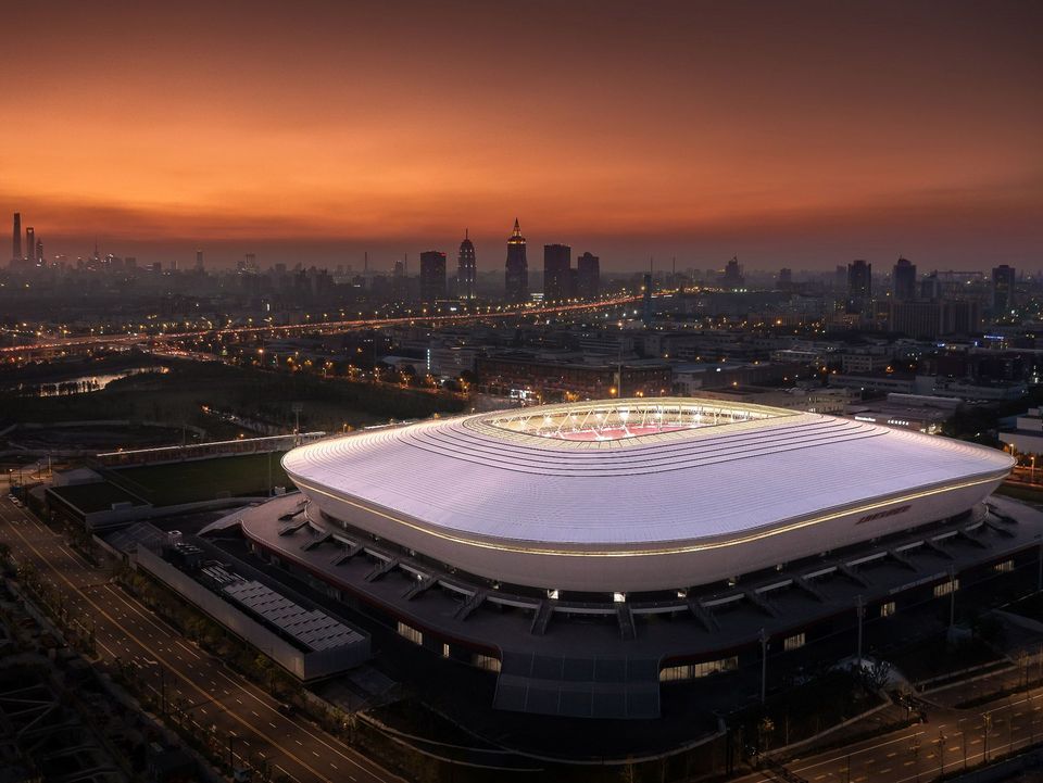 Abendliche Gesamtansicht eines Stadions als Luftbild mit leuchtender Dachkontur, ringförmiger Erschließung, angebundenen Parkflächen und Stadtsilhouette im Hintergrund.