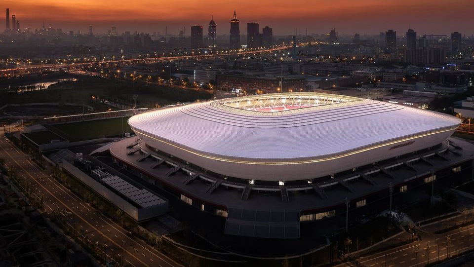 Abendliche Gesamtansicht eines Stadions als Luftbild mit leuchtender Dachkontur, ringförmiger Erschließung, angebundenen Parkflächen und Stadtsilhouette im Hintergrund.