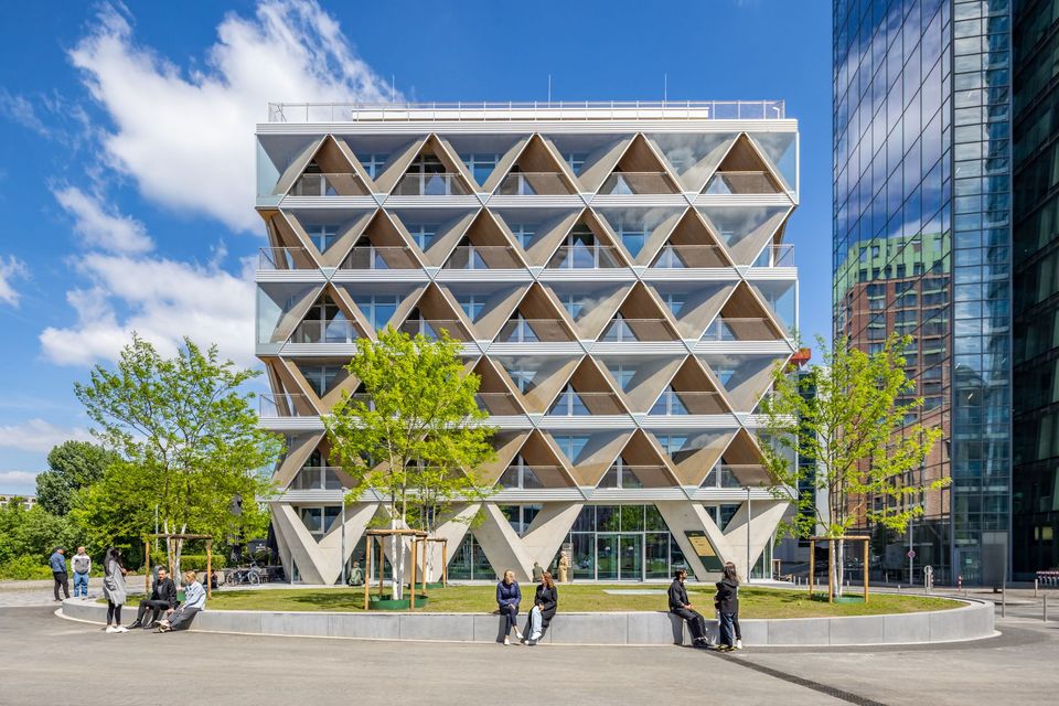 Frontal view of a multi‑storey timber‑hybrid building featuring a distinctive triangular façade pattern, regular balcony zones and an open forecourt.