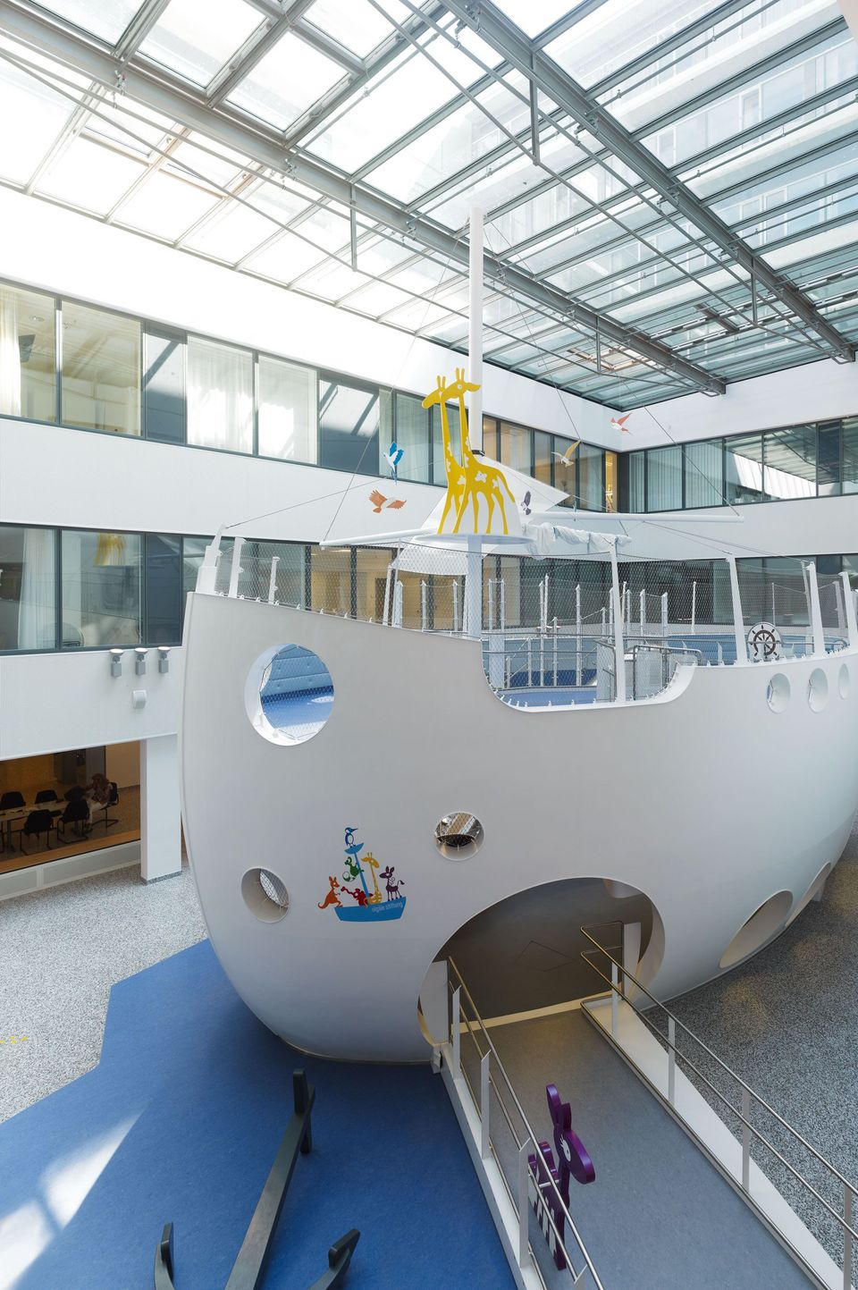 Spielbereich in Form eines großen weißen Schiffes mit Bullaugen und Mast, unter verglastem Dach im Atrium einer Klinik