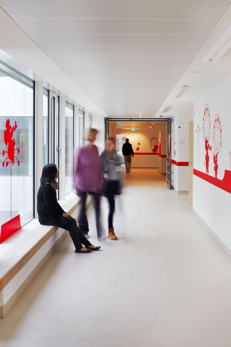 Heller Klinikflur mit großen Fenstern und roten Wandgrafiken, klare Linien und Sitzbank entlang der Glasfront