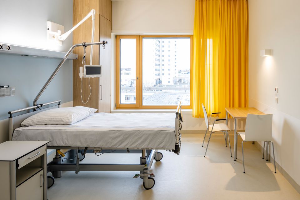 Innenaufnahme eines modernen Patientenzimmers mit Pflegebett, farbiger Akzentwand und großem Fenster mit gelbem Vorhang.