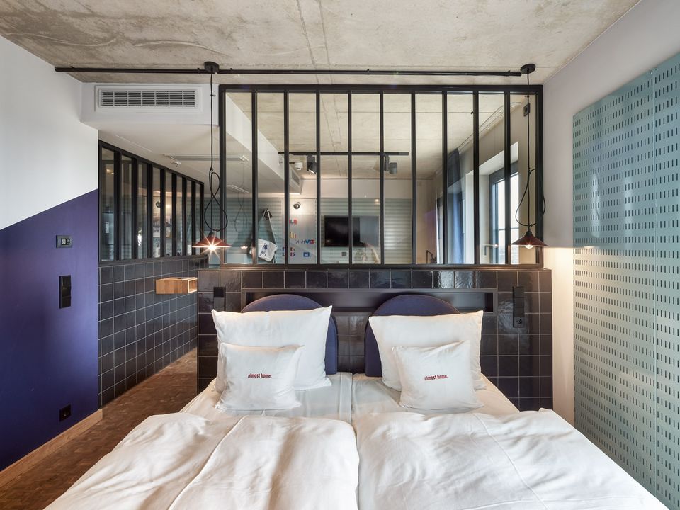 Hotelzimmer mit Doppelbett, dunkler Fliesenwand und industriellen Designelementen, offene Raumstruktur mit Glasabtrennung