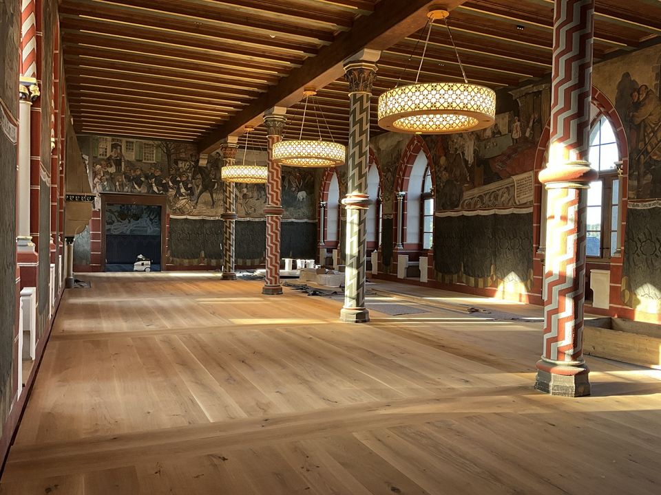 Innenaufnahme eines historischen Festsaals mit restaurierten Wandmalereien, dekorativen Säulen, hölzerner Deckenstruktur und neu verlegtem Boden im Zuge der Sanierung.
