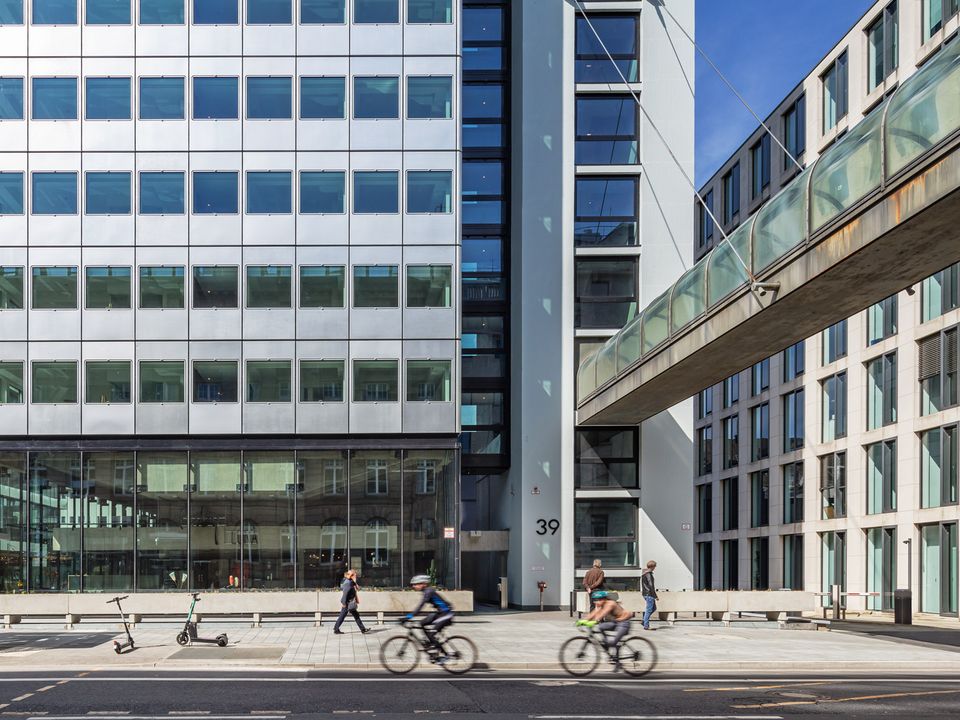 Frontansicht eines Hochhauses mit Glas- und Metallfassade, Fußgängerbrücke im Vordergrund, belebte Straße mit Radfahrern