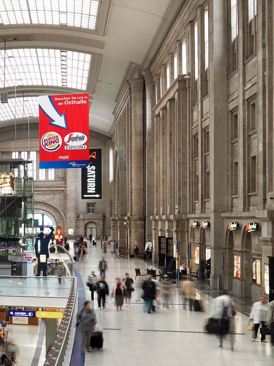 Innenaufnahme eines historischen Bahnhofs mit hoher Halle, sandsteinfarbener Fassadenarchitektur, großen Werbebannern und belebten Wegeachsen, die den Übergang zwischen Bahnsteigen und Shopping‑Bereichen zeigen.
