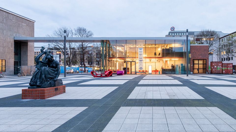 Außenaufnahme der Glasfoyer-Erweiterung mit durchgesteckter Transparenz zum Vorplatz und flankierenden Gebäuden.