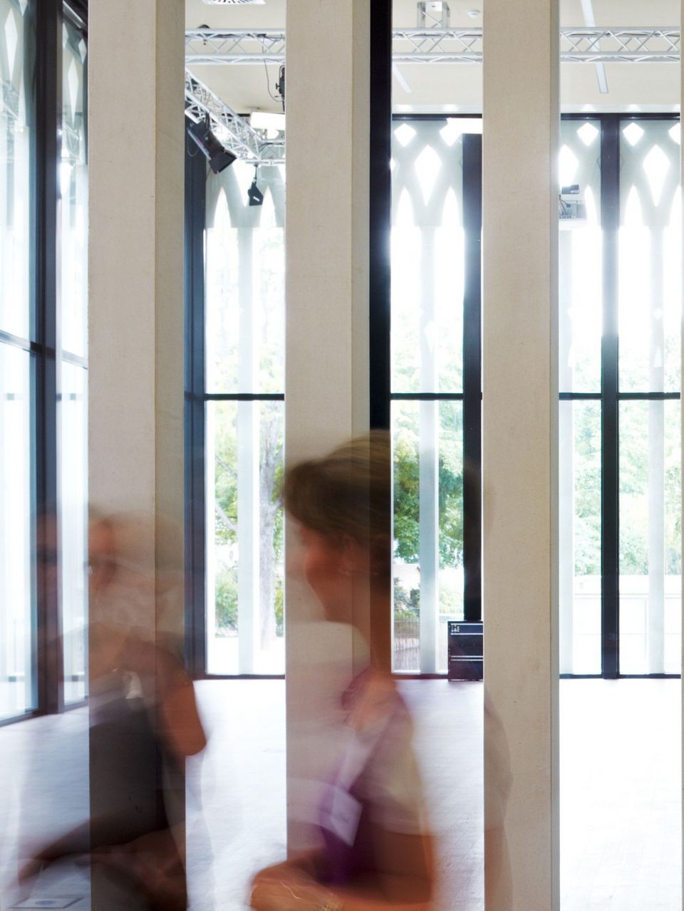 Innenaufnahme eines Raumes mit hohen weißen Pfeilern, Glasfassade und gleichmäßiger Tageslichtführung, die die vertikale Struktur und Transparenz der Architektur hervorheben.