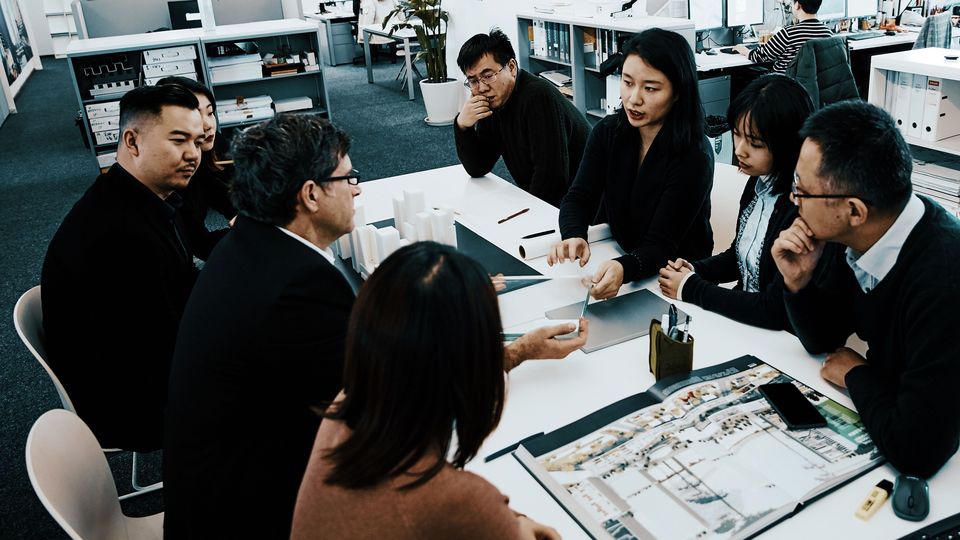 Innenaufnahme eines Architekturbüros mit sitzender Projektgruppe an einem Tisch, Modellen, Plänen und Arbeitsmaterialien in einem offenen Besprechungsbereich.