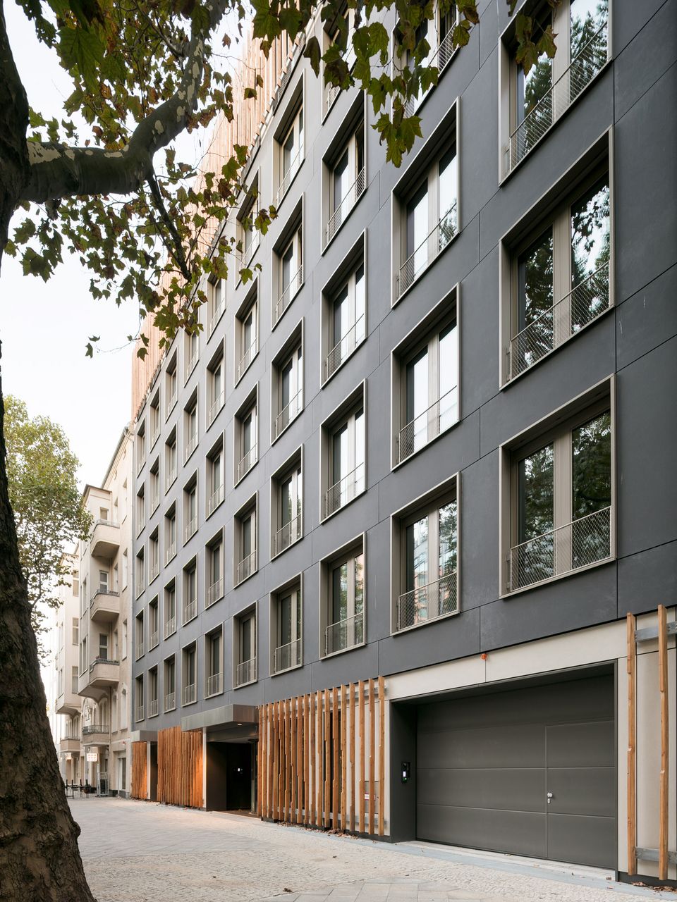 Straßenansicht eines modernen Wohn- und Bürogebäudes mit dunkler Fassade und Holzlamellen im Eingangsbereich
